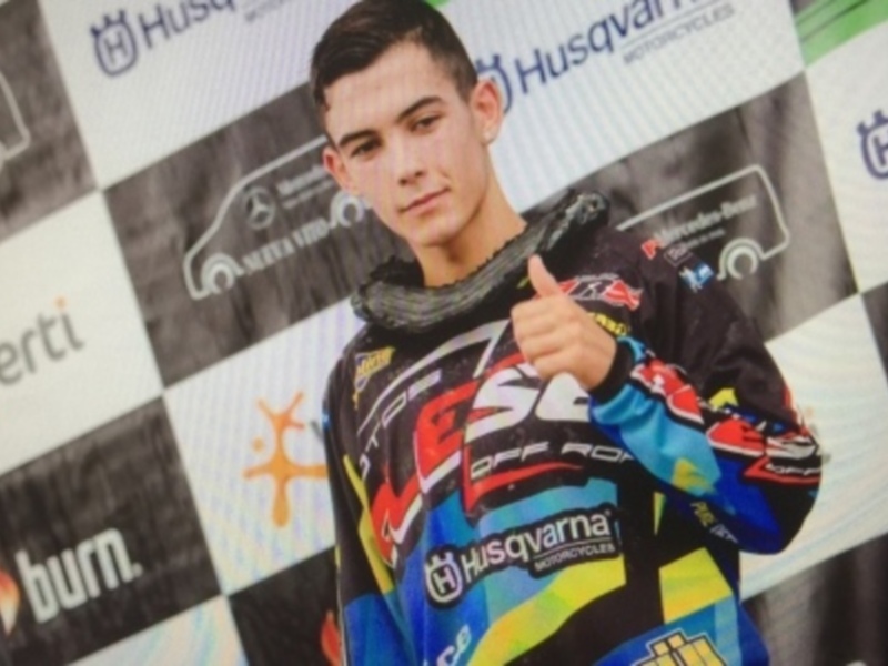 Un piloto de motociclismo muestra un gesto de satisfacción en un evento de carreras, con una camiseta con logos de sponsors en el fondo.