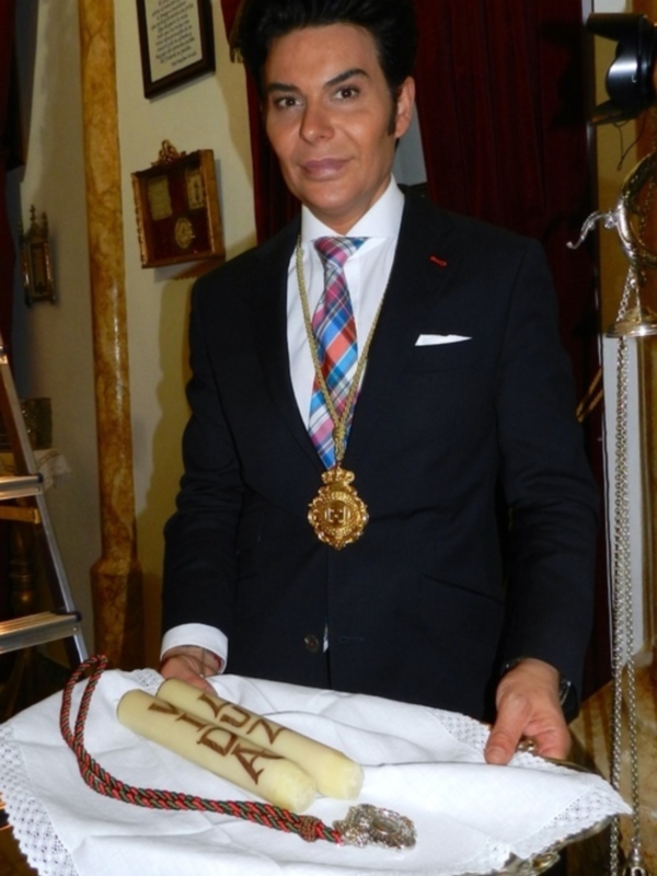 Un hombre en traje negro y corbata a cuadros, sosteniendo una bandeja con dos palitos de pan y una medalla dorada en la solapa.