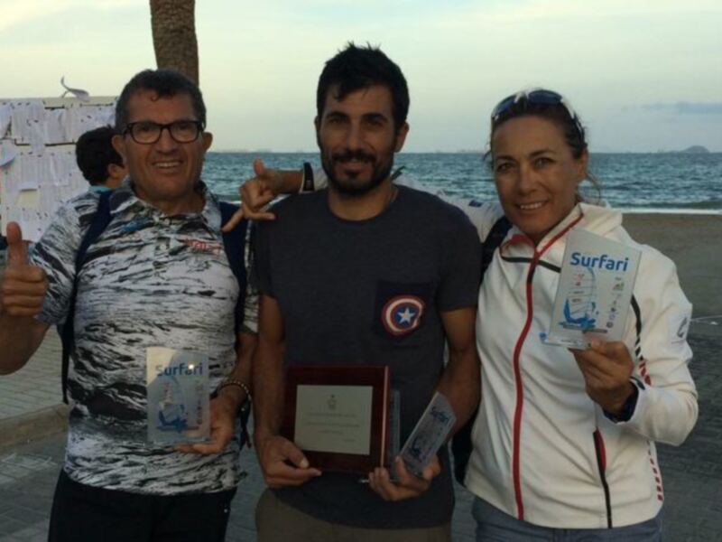 Tres personas posan con premios y una placa en una playa, celebrando Surfari.