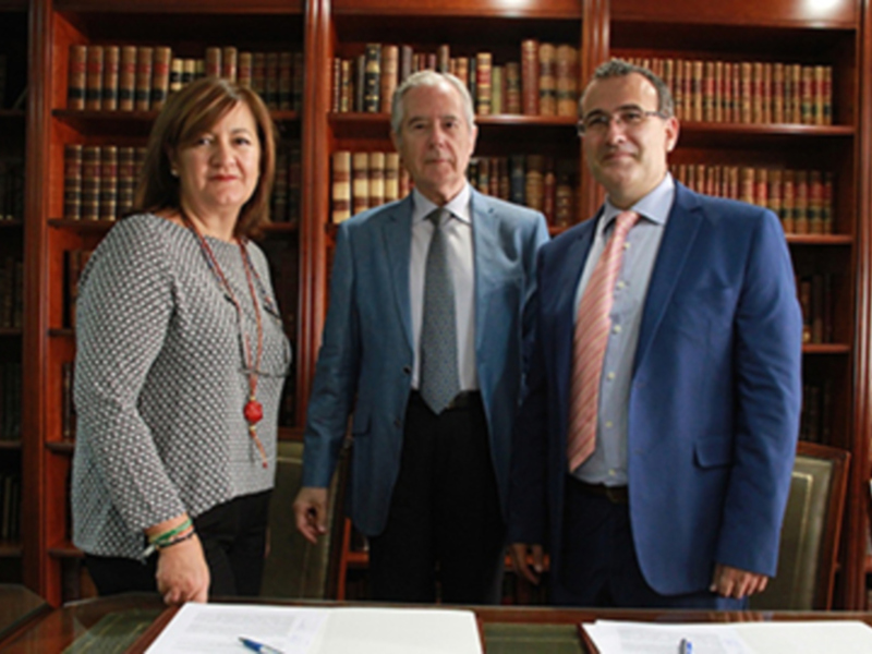 Tres personas posando tras una mesa con documentos, en un ambiente formal con estanterías de libros al fondo.