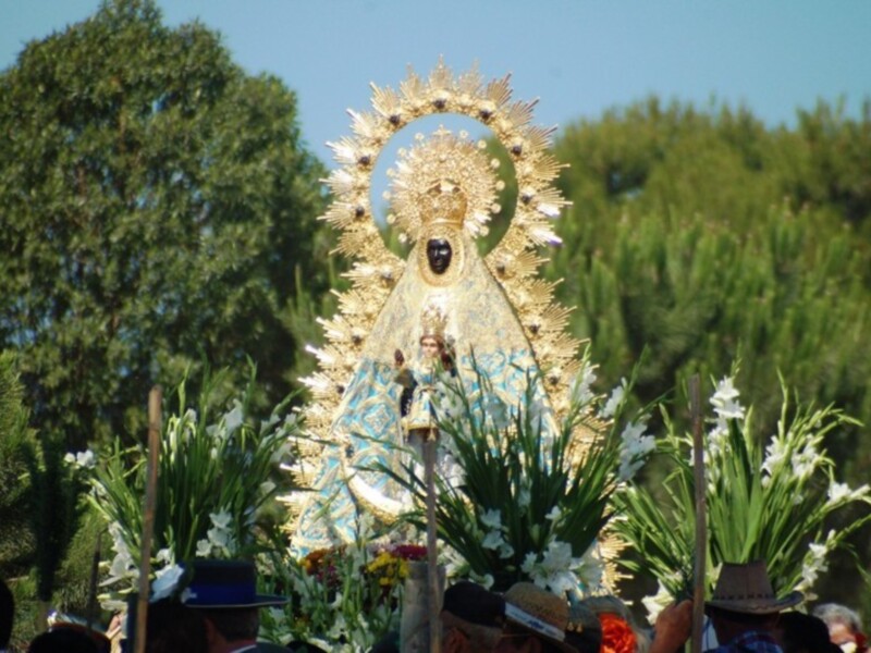 Procesión con imagen de la Virgen de la Cinta, adornada con flores y portada por devotos en un entorno natural.