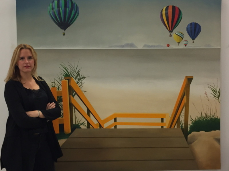 Una mujer posa frente a un mural que muestra un paisaje con un puente amarillo, palmeras y globos aerostáticos en el cielo.