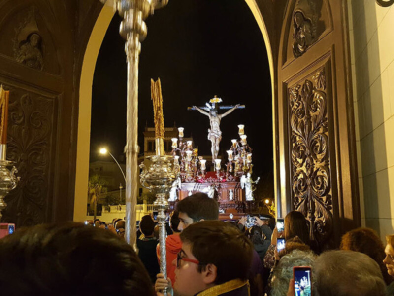 Procesión religiosa con estandartes y crucificado, iluminada por farolas y lámparas, en una noche de fiesta.