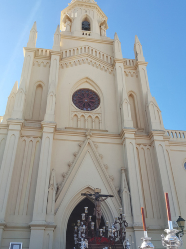 Procesión religiosa frente a una iglesia neogótica con un crucificado en el altar y fieles participando.