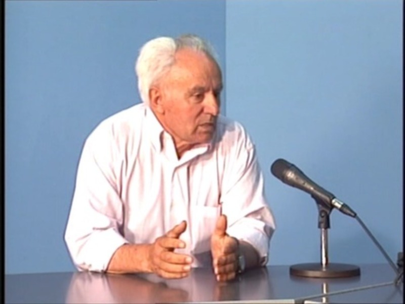Un hombre hablando en un evento, con un micrófono en frente de él y una pared azul en el fondo.