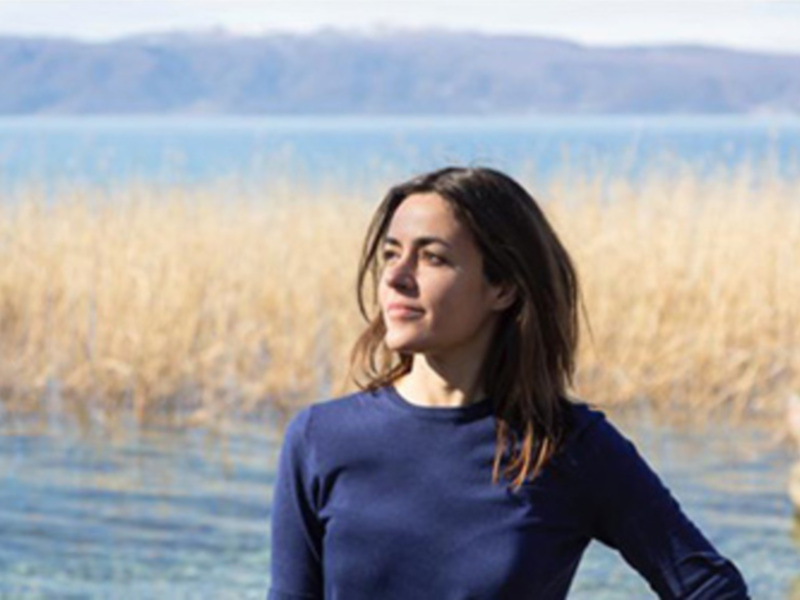 Una mujer con cabello largo y ondulado, vistiendo una camiseta azul, se encuentra frente a un lago con reedos en primer plano y montañas en el fondo. La escena transmite una sensación de tranquilidad y conexión con la naturaleza.