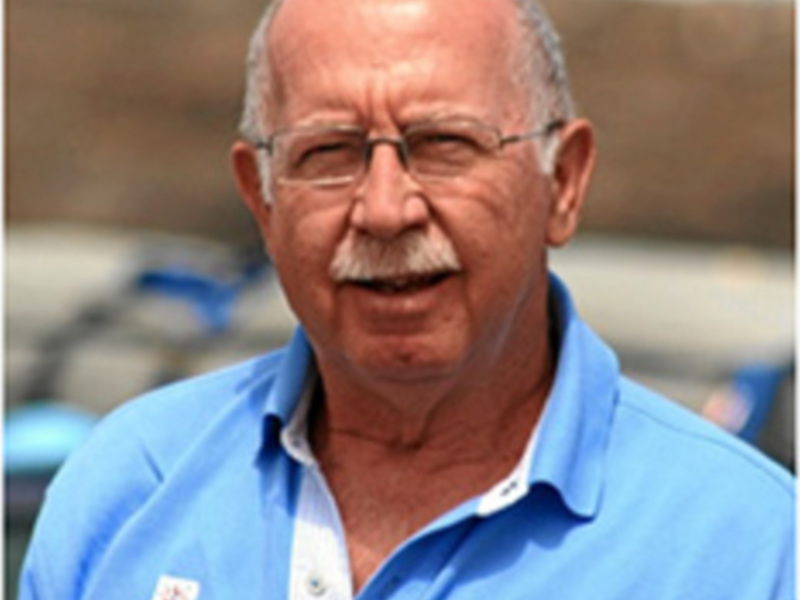 Un hombre con barba y gafas, vestido con una camisa azul, posa para la cámara. El fondo muestra un entorno urbano con estructuras de hormigón y una pista de carreras.