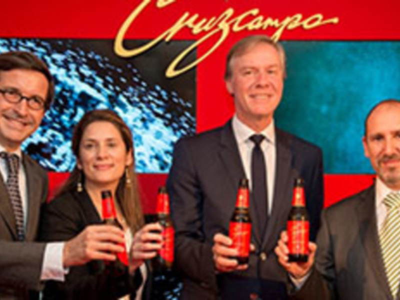 Cuatro personas sonrientes sostienen botellas de cerveza en una presentación de marca. El fondo es rojo con el logotipo de Cruzcampo.