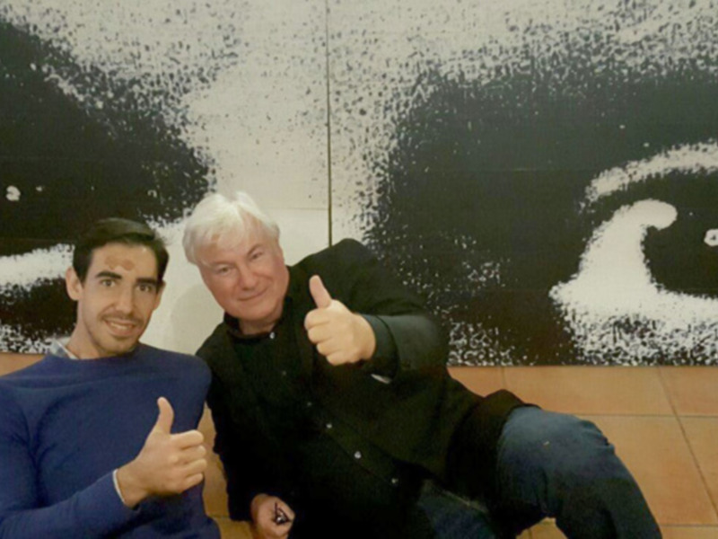 Dos personas sonrientes con los pulgares hacia arriba en frente de una obra de arte con un diseño de ojos en blanco y negro.