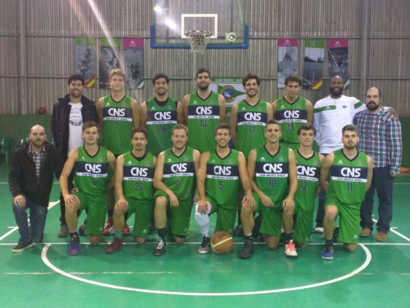 Equipo de baloncesto con camisetas verdes y la palabra 'CNS' en el pecho, posando en el interior de un gimnasio con una cancha de baloncesto visible.