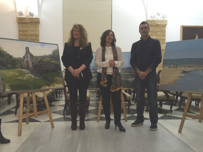 Exposición de pintura con tres personas posando frente a obras de arte en un espacio bien iluminado.