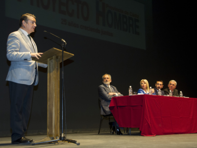 Presentación de un proyecto en la provincia de Cádiz, con un hombre hablando al micrófono y una mesa con cuatro personas escuchando. El texto en la pantalla dice 'Proyecto Hombre 25 años creciendo juntos'.