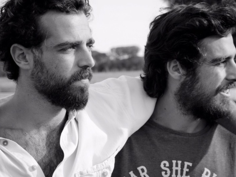 Dos hombres con barbas, uno con camisa blanca y el otro con camiseta oscura, posando juntos en un entorno al aire libre.