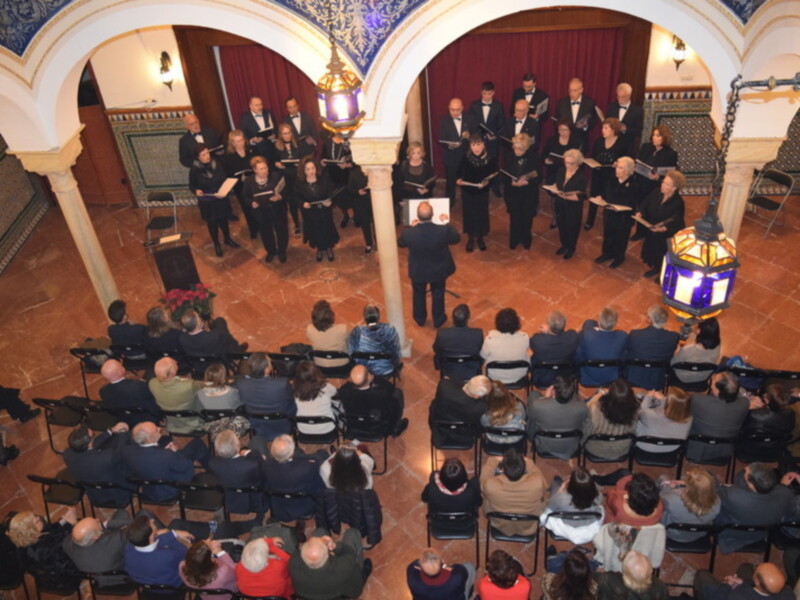 Concierto coral en un ambiente elegante y tradicional, con un coro en el centro y un público atento en las butacas.