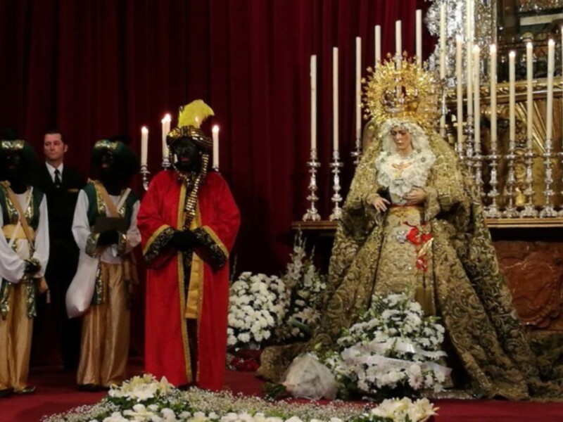 Fiesta religiosa con personajes vestidos con trajes tradicionales y una imagen de la Virgen en el centro, rodeada de flores y velas.
