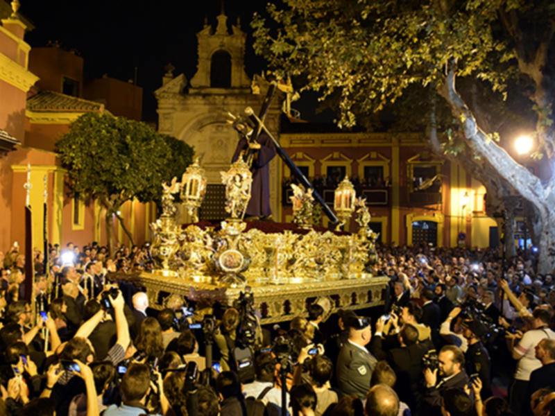 Fotografía nocturna de una procesión religiosa en una ciudad, con una imagen dorada y cristalina en el centro, rodeada por una multitud de personas que la observan con atención, iluminada por faroles y luces de la ciudad.