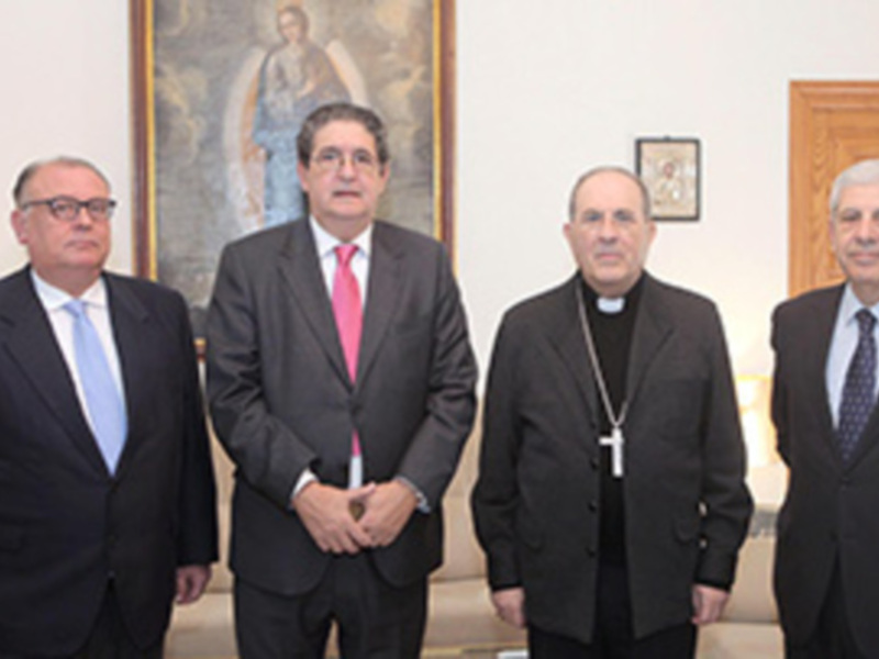 Tres personas en trajes, una con un círculo en la camisa, posan frente a una imagen religiosa. La escena parece ser formal, posiblemente en un entorno institucional o religioso.
