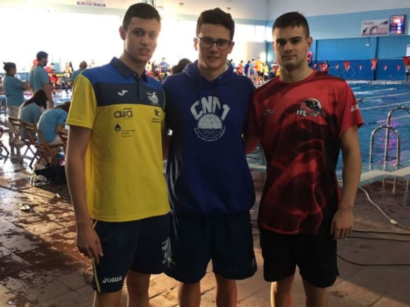 Tres jóvenes posan juntos en una piscina cubierta, vestidos con ropa deportiva de colores vibrantes, con una competencia o evento en el fondo.