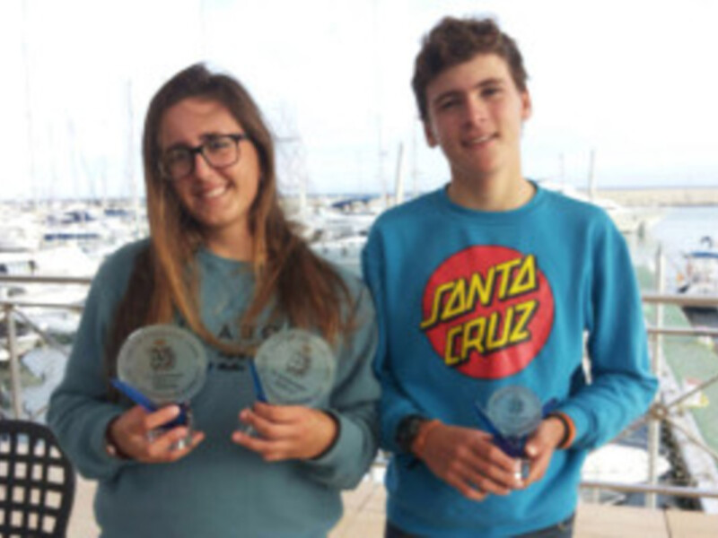 Dos jóvenes sonrientes posan con medallas en un puerto marítimo.