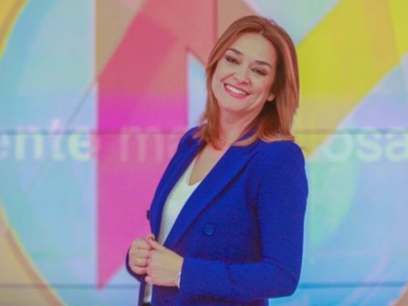 Una presentadora de televisión sonríe ampliamente frente a un fondo de colores brillantes, con una sonrisa radiante que destaca su alegría y profesionalidad.