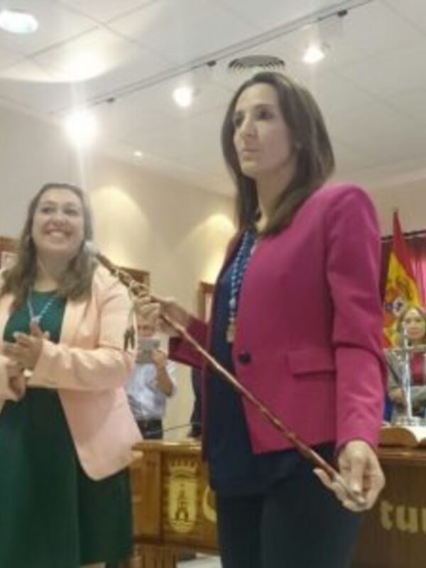 Mujeres en una reunión oficial con banderas y un diploma en el fondo.