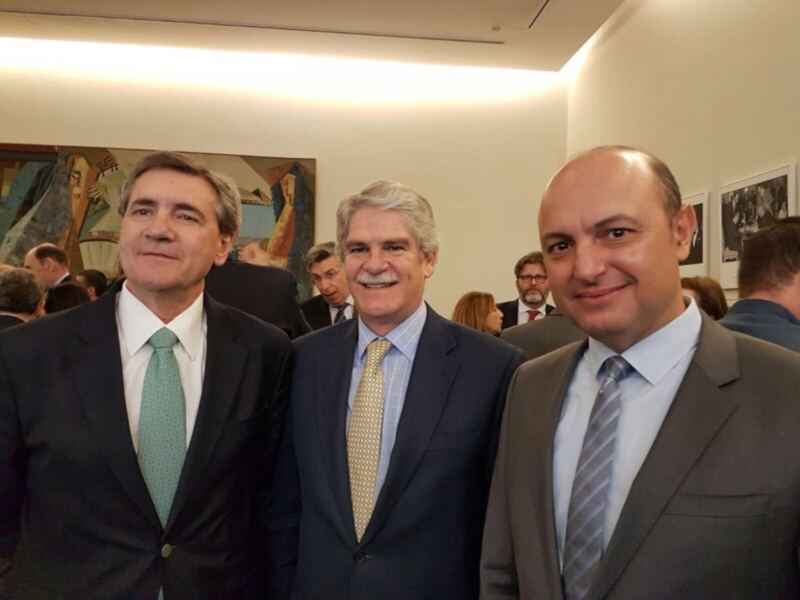 Tres hombres en trajes sonrientes en un evento formal, fondo con arte y personas en el fondo.