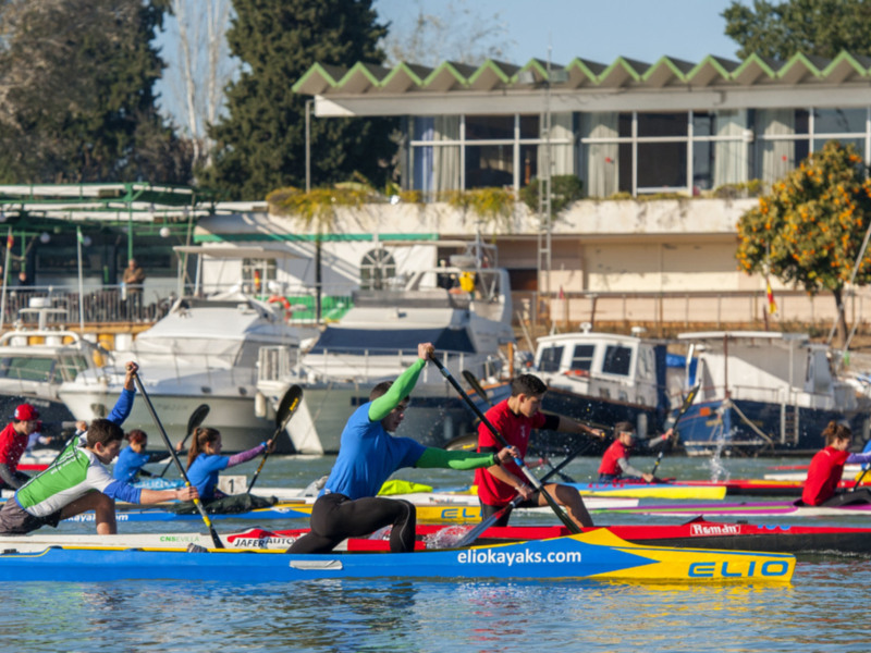Equipo de kayak en competición en un río con barcos y edificios al fondo.