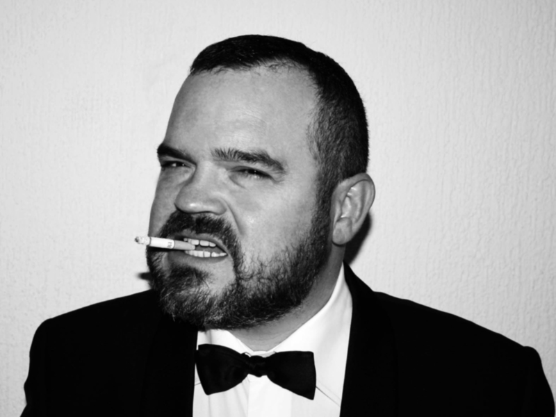Un hombre en traje negro y corbata sostiene una cigarrilla en la boca, con una expresión seria y pensativa.