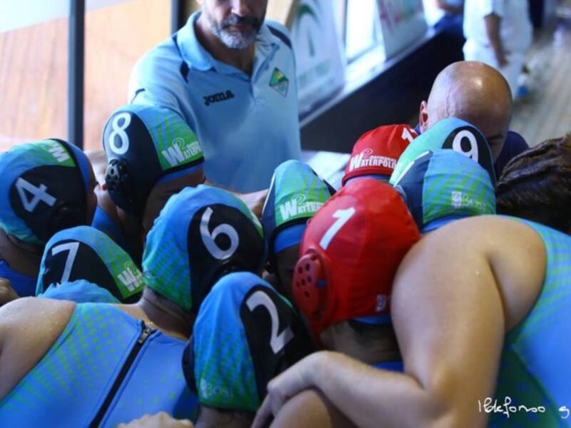 Equipo de waterpolo reunido en una conferencia de prensa, con los jugadores vestidos con cascos numerados y el entrenador de pie detrás de ellos.