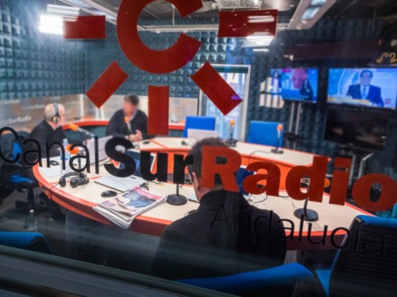 Sala de radio con dos personas sentadas en una mesa redonda, con micros y pantallas en el fondo.
