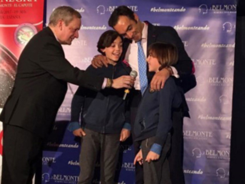 Persona adulta y dos niños en un evento formal, uno de los adultos entrega un micrófono a uno de los niños.