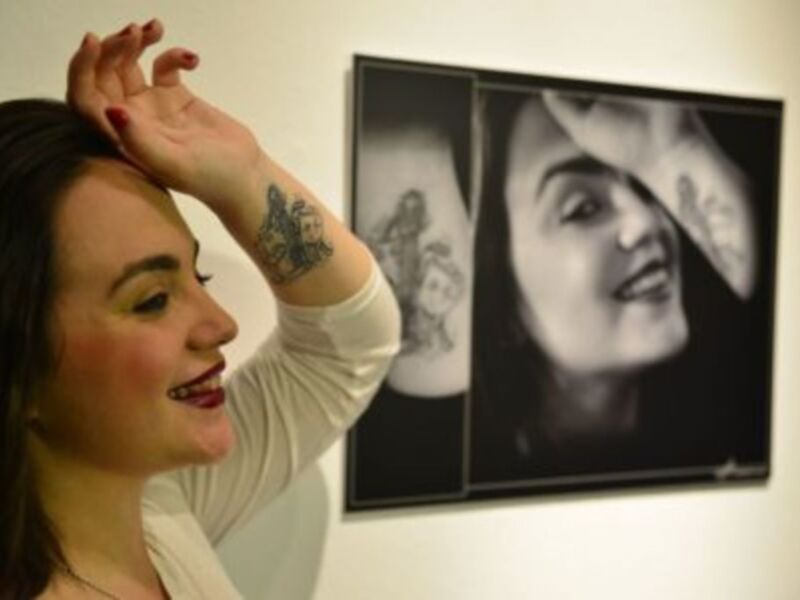 Una mujer sonríe junto a una fotografía en blanco y negro de una persona en una exposición de arte.