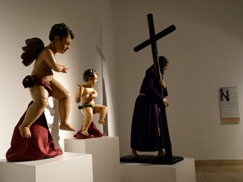 Esculturas de figuras infantiles y una figura adulta portando una cruz en una exposición.