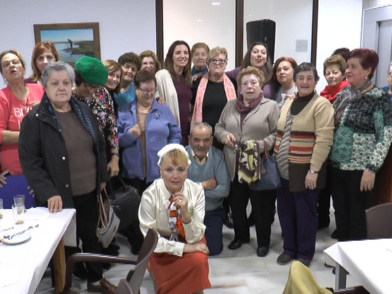 Grupo de mujeres en una reunión social o evento, sonrientes y reunidas en un salón con mesas y sillas.