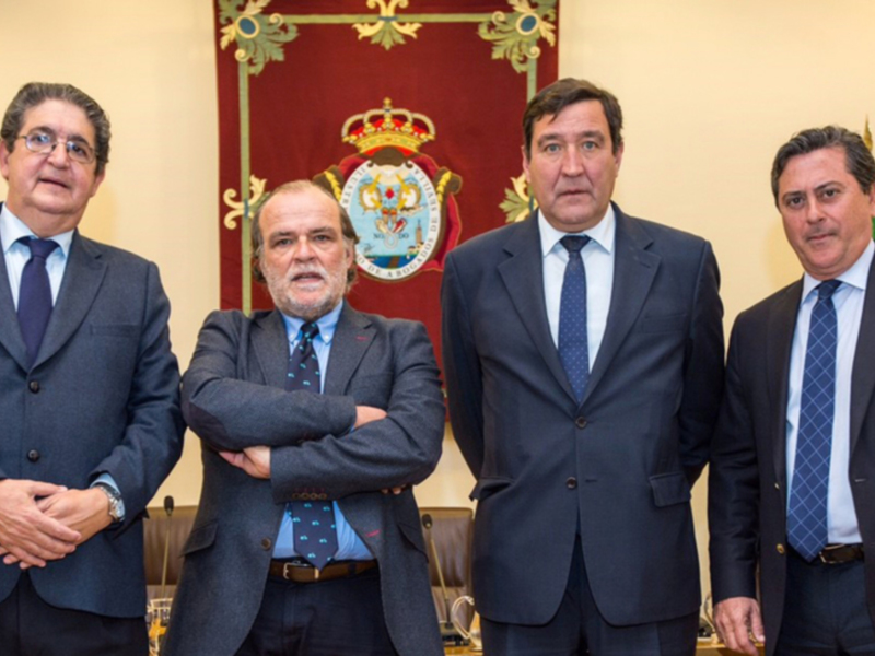 Tres hombres en trajes posan frente a una bandera española y un escudo heráldico, posiblemente en una reunión oficial.