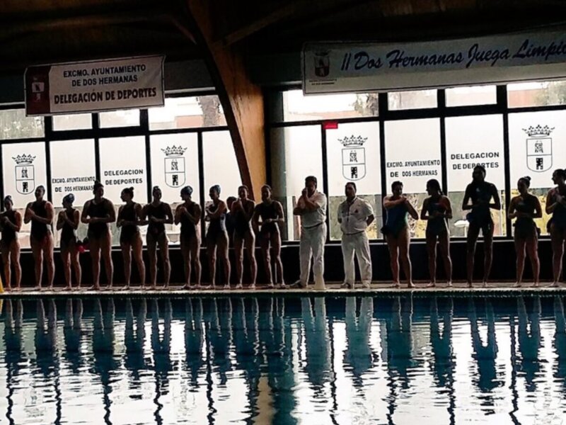 Equipo de natación en una piscina, listo para competir. Detalle de la piscina y el edificio de fondo con letreros de la delegación de deportes y el Ayuntamiento de Dos Hermanas.