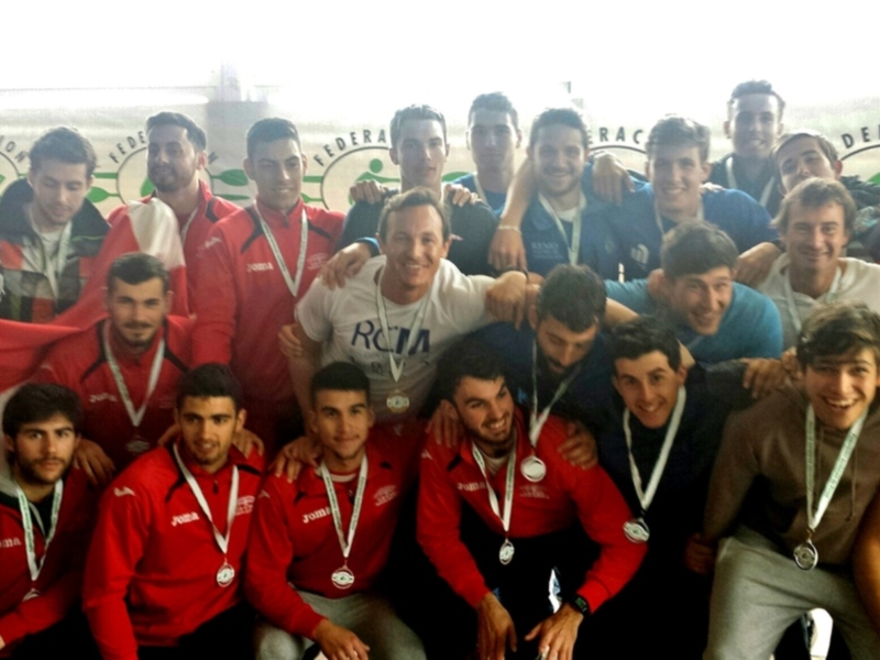 Un equipo de atletas celebrando su victoria, todos sonrientes y con medallas en el cuello. La bandera nacional se levanta en el fondo, mostrando orgullo y unidad.