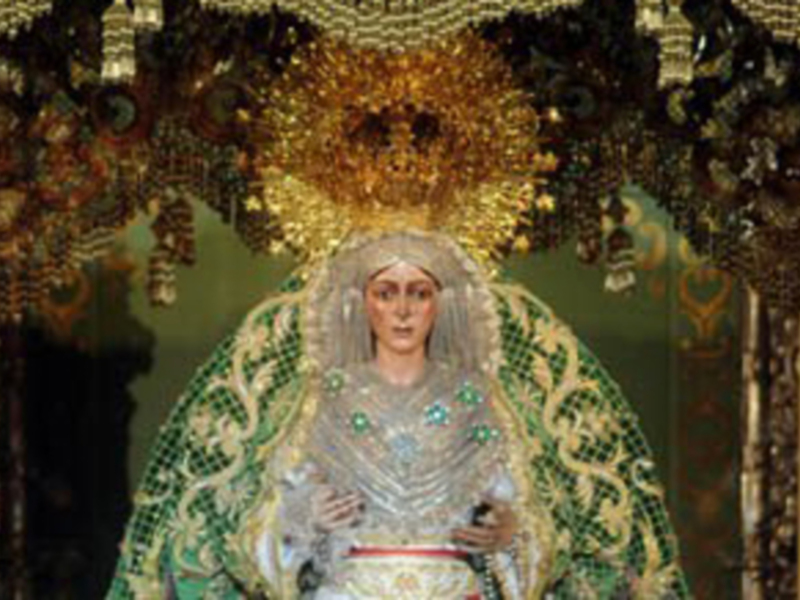 Imagen religiosa de una Virgen María vestida con joyas y una corona dorada, en un entorno decorado con luces y adornos.