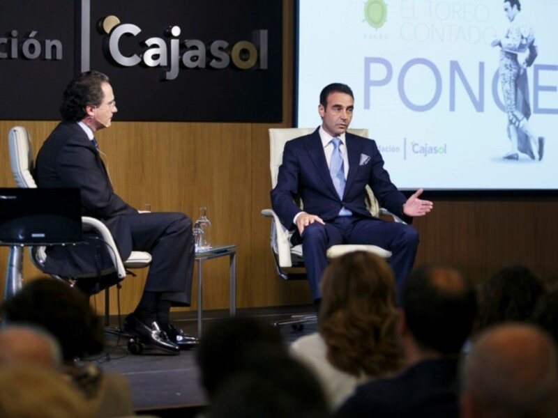 Dos hombres en trajes se sientan en sillas frente a una pantalla con la palabra 'Cajasol' y 'PONCE'. La audiencia se puede ver en el fondo.