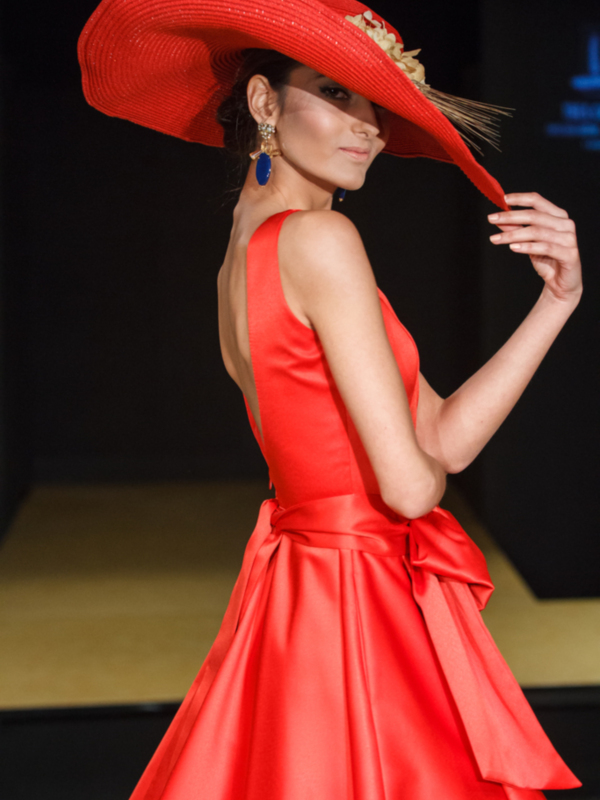 Modela con elegancia en un desfile de moda, luciendo un vestido rojo con un gran sombrero y detalles en azul.