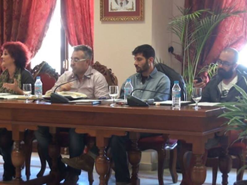 Un grupo de personas se reúnen en una sala con cortinas rojas y plantas verdes, sosteniendo documentos y charlando en un ambiente formal.