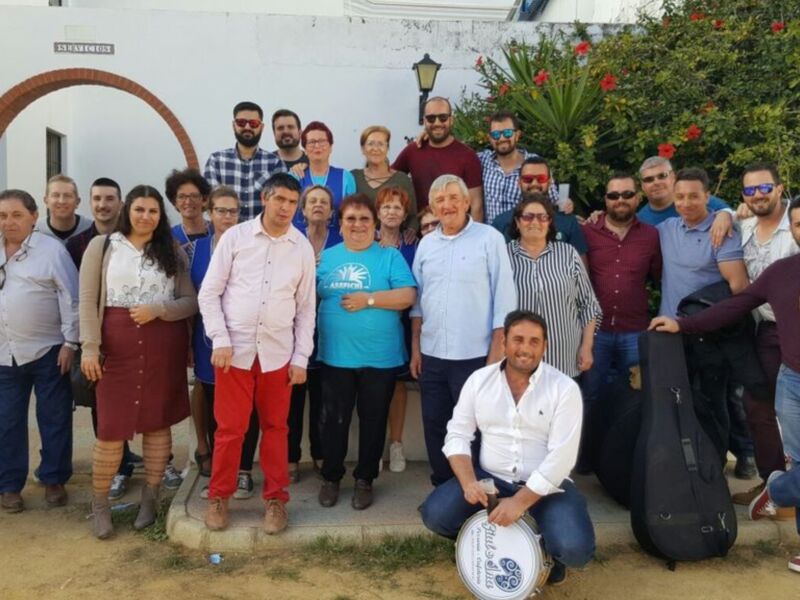 Grupo de personas reunidos en una foto de grupo al aire libre, posando frente a una fachada blanca con detalles arquitectónicos y plantas en el fondo.