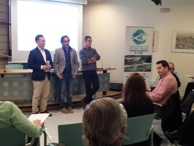 Una conferencia con tres hombres en el escenario y varios asistentes sentados en el público, con una pantalla de proyección en el fondo. El ambiente parece ser profesional y educativo.