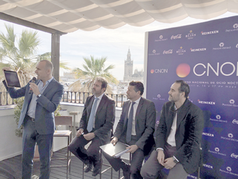 Un evento de negocios con un hablante en un escenario al aire libre, destacando la torre de Sevilla en el fondo.