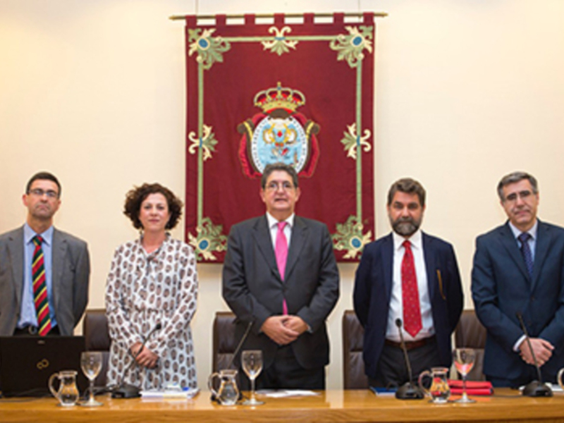 Una reunión formal en un salón de conferencias, con cuatro personas en trajes y dos banderas de España y Andalucía en el fondo.