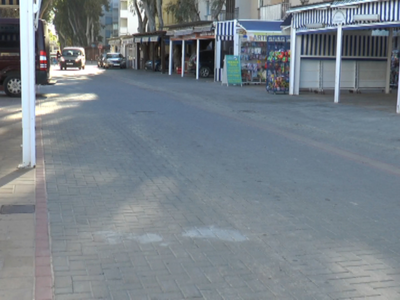 Vista de una calle vacía con tiendas y sombras de árboles en un día soleado.