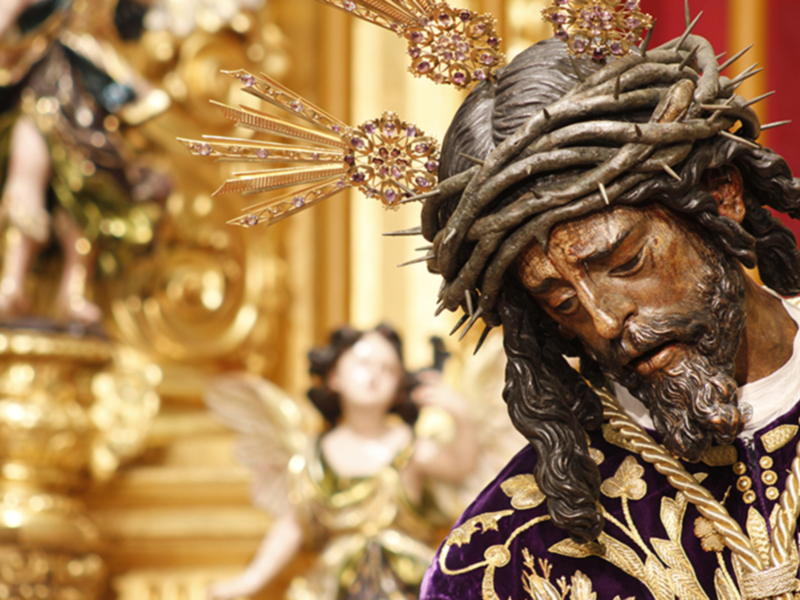 Figura religiosa con corona de espinas y vestimenta ornada, en un entorno dorado con detalles dorados y angelotes al fondo.