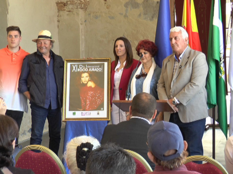 Presentación del XI Festival Internacional Rocío Jurado en un evento oficial con banderas y un panel con su imagen.
