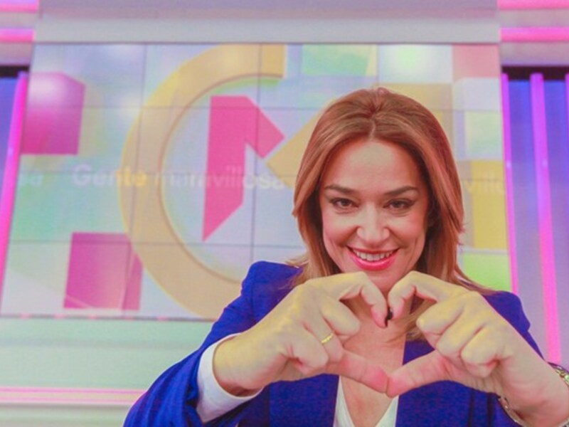 Una mujer sonríe y hace un corazón con las manos en un estudio de televisión con un fondo colorido.