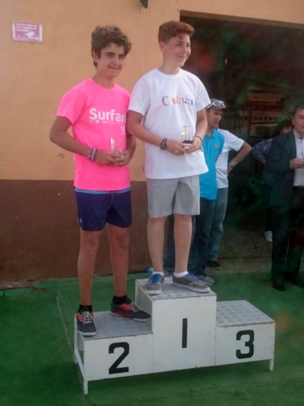 Dos jóvenes en un podio, uno con camiseta rosa y otro con blanco, sonríen mientras sostienen trofeos. A la derecha, hay personas observando el evento. Detalle de una pared con una placa roja en el fondo.
